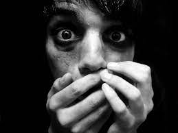 A man in a dimly lit room, covering his mouth with his hands, expressing shock or secrecy.