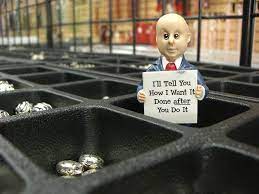 A tiny man holds a sign inside a box, showcasing his message, which reads "I'll tell you how I want it done after you do it."