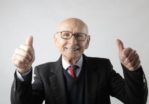 An older man in a suit and glasses, smiling and showing approval with a thumbs up gesture.