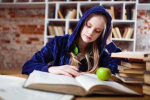 A girl in a hoodie engrossed in an open book, holding an apple.
