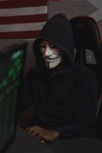 A man in a mask and hoodie sitting at a computer desk.