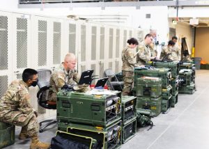 Army soldiers working on laptops in a computer-filled room, collaborating on tasks and utilizing technology.