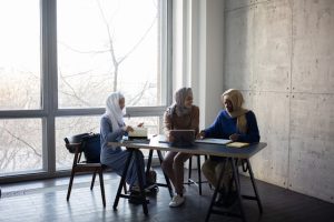 Three women sitting at a table near a window, engaged in conversation and enjoying each other's company.