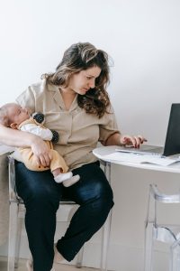 A woman multitasking at her desk with a baby and laptop, balancing work and motherhood simultaneously.